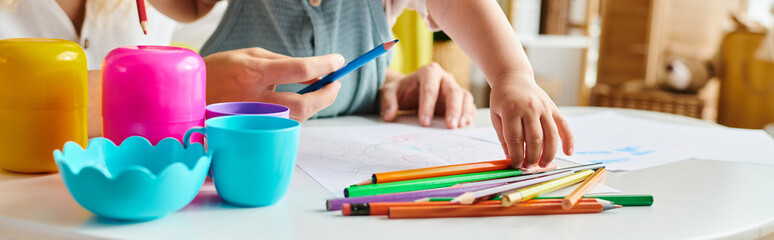 Wall Mural - A woman sits at a table, focused on a bunch of crayons with her toddler daughter, embracing the Montessori method of education.