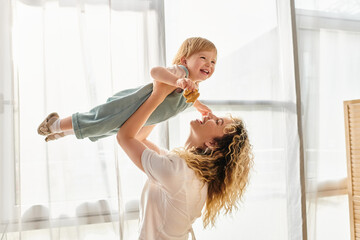 a curly mother joyfully lifts her toddler daughter up into the air, expressing love and playfulness 