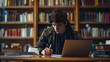 © nopommajun - A young individual is taking notes next to a laptop in a library, with bookshelves in the background