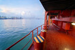 © Image Source - The image captures a vibrant view from the deck of a ship showing red and yellow railings, calm sea water, and container cranes at a port in the distance under a partly cloudy sky.