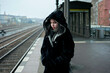 © Connect Images - A young adult woman stands on a train platform, wearing a warm black coat with a hood, looking to the side with a slightly open mouth, conveying a sense of anticipation or contemplation.