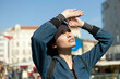 © Connect Images - A young woman is outdoors on a sunny day, shading her eyes with her hand as she looks upward, with a city street and buildings in the background.