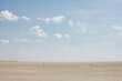 © Image Source - The image showcases a vast, sandy beach with a calm blue sky overhead dotted with white clouds. People appear as tiny figures in the distance, emphasizing the expansive nature of the beach setting.