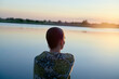 © Image Source - A young woman is sitting by a tranquil lake at sunset, gazing at the serene water reflecting the soft hues of the twilight sky.