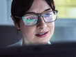 © Connect Images - A woman with glasses reflecting colorful data graphs is looking at a computer monitor with a focused expression.