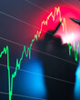 © Image Source - A blurred silhouette of a person is seen in the foreground with a background of a stock market chart displaying a fluctuating trend line in red and green.