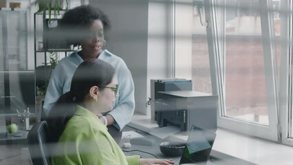 Wall Mural - tilt up medium shot of two young multiethnic women looking at laptop screen and talking while co-wor