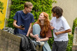 © Connect Images - Three teenagers are socializing outdoors, with one showing something on a mobile phone to a smiling girl while another stands by with a beverage in hand.