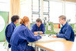© Image Source - Four individuals in blue work attire are engaged in a discussion around a table in an industrial setting, with machinery in the background.