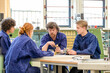 © Connect Images - Four individuals in blue work attire are engaged in a discussion around a table in a workshop setting with machinery in the background.
