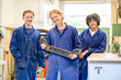 © Connect Images - Three apprentices stand side by side dressed in blue work overalls in a workshop, smiling as they hold a large metallic tool together.