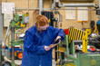 © Connect Images - A focused individual with red hair, wearing a blue coverall, is inspecting a document while standing next to an industrial machine in a workshop.