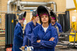 © Connect Images - A confident young  woman with curled hair wearing a welding helmet and a blue work suit stands with arms crossed in a workshop setting