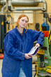 © Connect Images - A young woman with red hair, wearing a blue coverall, stands in an industrial setting, holding papers and leaning on machinery.