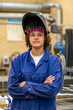© Connect Images - A confident young worker is standing with arms crossed, wearing a welding helmet flipped up, and dressed in a blue protective work suit against an industrial backdrop.