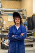 © Connect Images - A confident young welder stands with crossed arms, wearing a blue overcoat and a protective welding helmet flipped up on their head in a workshop setting.