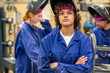 © Connect Images - A young person with curly hair stands confidently in a welding workshop, wearing a blue safety uniform and helmet, with focused classmates in the background.
