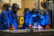 © Connect Images - Three individuals are engaged in welding activities at a workshop, wearing protective gear including darkened helmets and blue aprons.