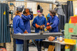© Connect Images - Four individuals in blue work uniforms and protective welding helmets are focused on a task at a workstation with various tools and a metal piece.
