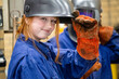 © Connect Images - A young female welder in a blue coverall lifts her welding helmet with a gloved hand, smiling at the camera in an industrial setting.