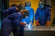 © Connect Images - Four individuals are engaged in welding work at a metal workshop, wearing protective gear including masks and gloves.