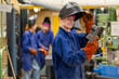 © Connect Images - A smiling worker in protective gear holding a tool in an industrial environment with colleagues in the background.