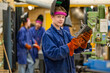 © Connect Images - A young male worker wearing a blue work uniform and orange gloves holds a metal piece in a workshop with machinery and colleagues in the background.