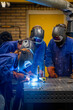© Connect Images - Three individuals are focused on welding metal in a workshop environment, wearing protective gear, including welding helmets and gloves, with bright welding sparks visible.
