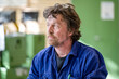 © Connect Images - A man with curly hair wearing a blue work uniform is looking to the side with a reflective expression, indoors, likely in a workshop environment.