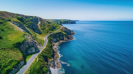 Wall Mural - Overhead view of a winding coastal road with vibrant green cliffs on one side and a deep blue sea on the other, under a clear azure sky.