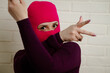 © Anton Dios - A woman wearing a bright pink balaclava dances energetically, striking unique poses with her hands against a clean, white brick wall background