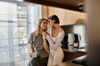 © Bliss - Young lesbian couple standing in a stylish kitchen inside a hotel room, holding coffee cups and chatting.