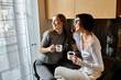 © Bliss - Young lesbian couple enjoy morning coffee together while sitting on window sill inside hotel room.