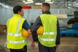 © BESTIMAGE - Two male factory workers wearing yellow safety vests walk and talk in a large industrial building.