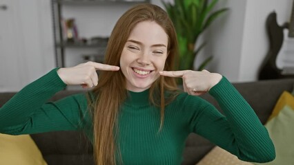 Wall Mural - Cheerful young blonde woman, in cozy home, pointing to her perfect white teeth. sporting her sweater, her joyous smile spotlights great dental health!