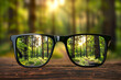 © Mikhail - A black-framed glasses focuses on a vibrant forest landscape, set against a blurred background of greenery. Shallow depth of field