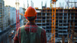 © Aleksandra - A construction worker in a hard hat standing with their back to the camera, looking at a large construction site with cranes and scaffolding,showcasing responsibility and oversight in the construction