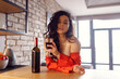 © Nataliya - Sexy young woman holding glass with wine, leaning at the table in the room at home, wearing red silky pajamas, looking at camera. Rest time concept