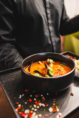 Wall Mural - Close-up of a bowl of spicy seafood soup garnished with herbs, presented by a person in a dark attire, against a blurred background