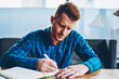 © BullRun - Skilled red haired student writing down homework in copybook studying at wooden table in coworking space.Pensive young man dressed in casual shirt noting checklist in notepad during free time