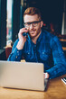 © BullRun - Portrait of successful student in eyeglasses talking on smartphone with friend while searching information on websites on laptop device during e-learning in stylish coffee shop interior using free 4G