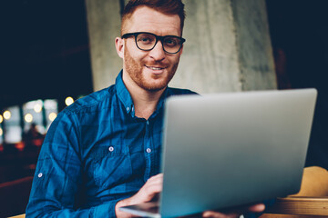 positive bearded male graphic designer with red hair and eyeglasses smiling at camera while working on new project at modern laptop computer connected to wireless 4G internet in coffee shop