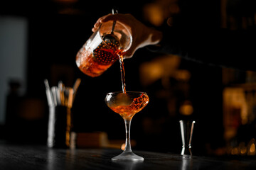 Bartender's hands pouring cocktail from mixing glass, straining it through a strainer into a stemmed glass