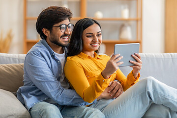 Wall Mural - Indian man and woman are sitting on a couch, engaged with a tablet in their hands. They are focused on the screen, possibly watching a video or browsing the internet together