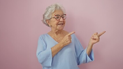 Wall Mural - Cheerful, mature woman with grey hair, standing and pointing to the side, inviting you to check out her favourite product! come join her over the vibrant pink background.