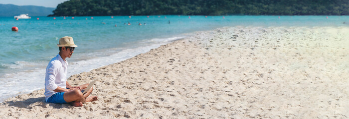 Wall Mural - Banner Man traveler use laptop on summer beach blue sky. Asian man travel relax sitting summer beach island. Man typing laptop computer holiday business trip freelance working online with copy space