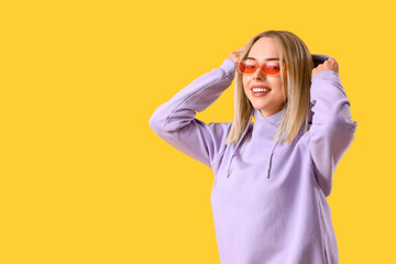 Poster - Beautiful young woman in stylish hoodie and sunglasses on yellow background