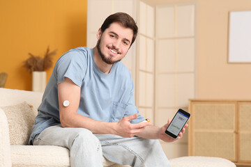 Wall Mural - Diabetic young man with glucose sensor, glucometer and mobile phone sitting on sofa at home