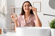 © Pixel-Shot - Young woman in pajamas with cotton pad and micellar water for removing makeup near sink at home