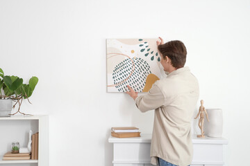 Young man hanging painting on light wall at home, back view